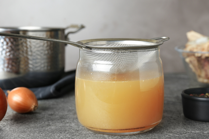 Clear jar with pork pigs feet bone broth liquid and metal strainer on a gray surface with onions and a pot in the background.