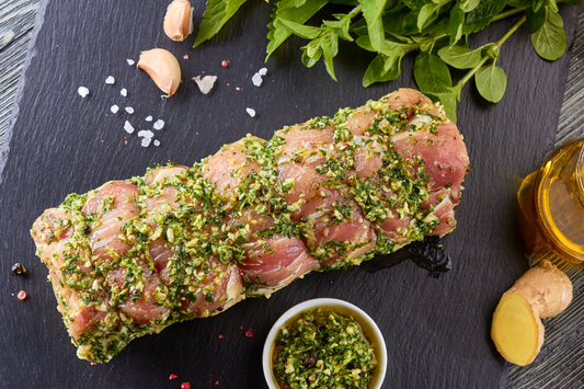 Marinated local farm pastured pork tenderloin with herbs on a dark surface, surrounded by spices and a bowl of marinade.