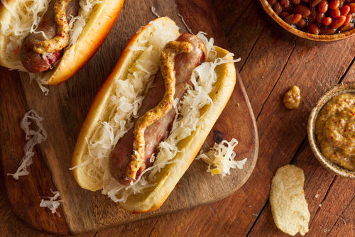 A non-GMO local forest raised pork bratwurst sausage in a bun with sauerkraut and mustard, displayed on a wooden table.