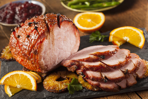 A carved local pastured pork smoked ham on a serving platter with slices of orange and herbs, alongside a bowl of cranberry sauce and a dish of green beans. The farm-raised holiday ham has a golden-brown color.
