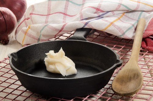 A small amount of solidified pasture raised pork lard in a black cast iron skillet with a wooden spoon beside it, on a kitchen towel with a pattern of red and white. There are onions in the background.