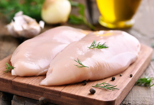 Raw boneless skinless pasture-raised non GMO chicken breasts on a wooden cutting board, garnished with herbs from our local chicken delivery in NC Triangle.