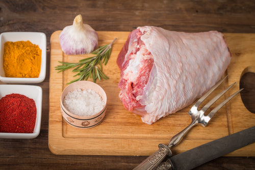 A raw non-GMO pasture-raised turkey thigh on a wooden cutting board with a knife, salt, pepper, thyme, and garlic on the side.